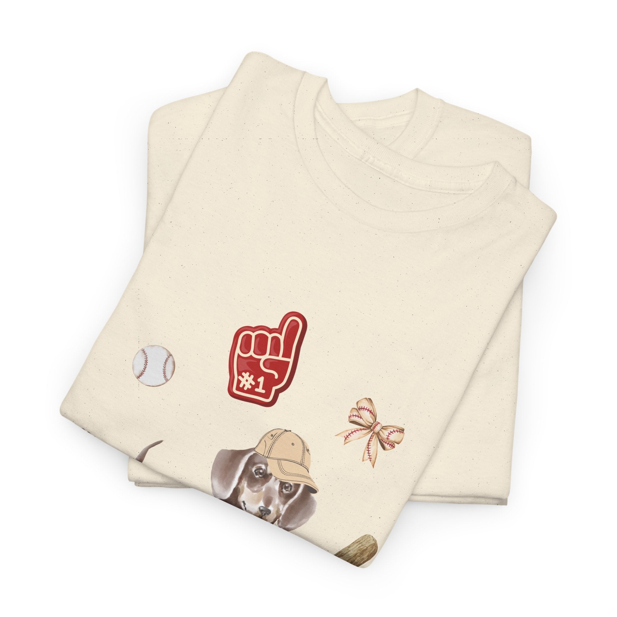 A folded beige t-shirt featuring a graphic design of a dog in a baseball hat surrounded by a circle of balls, bats, and helmets.