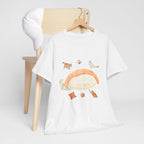 A white t-shirt hanging on a chair, featuring a graphic design of a piece of nigiri sushi surrounded by a circle of cats, yarn balls, and cushions.