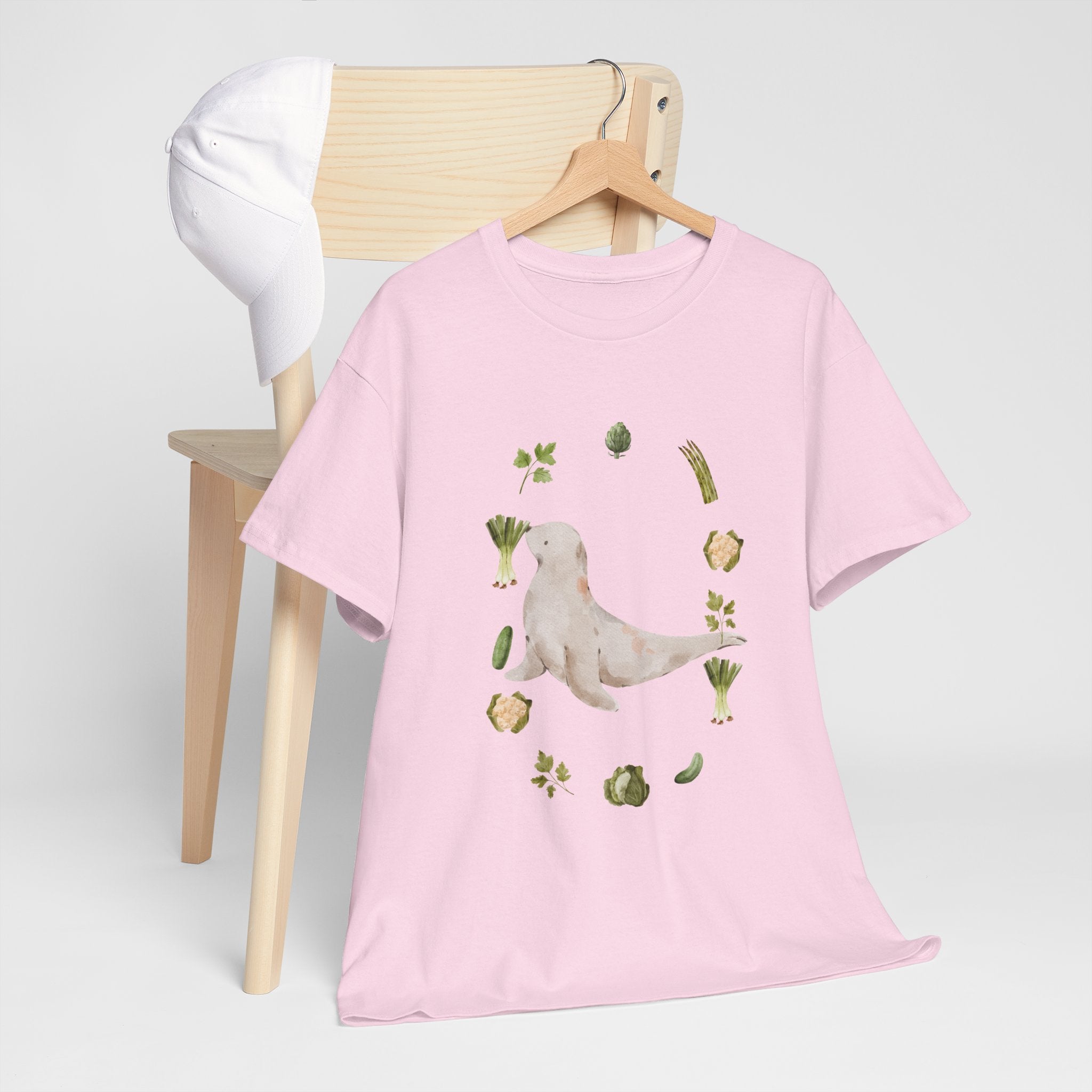 A pink t-shirt hanging on a chair, featuring a graphic design of a sea lion surrounded by green onions, cabbage, and broccoli.