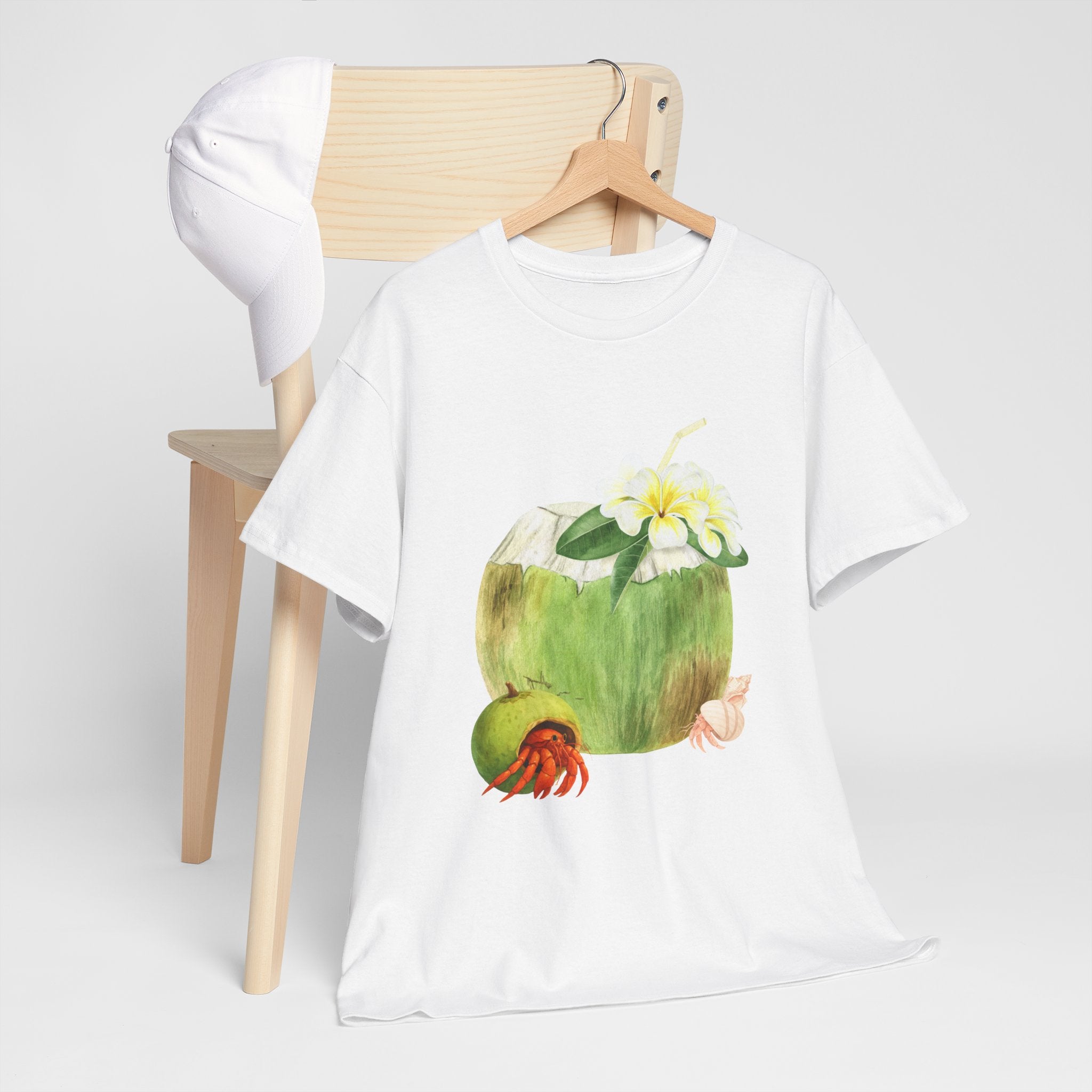 A white t-shirt hanging on a chair, featuring a graphic design of a hermit crab in a coconut shell and a straw-topped coconut.