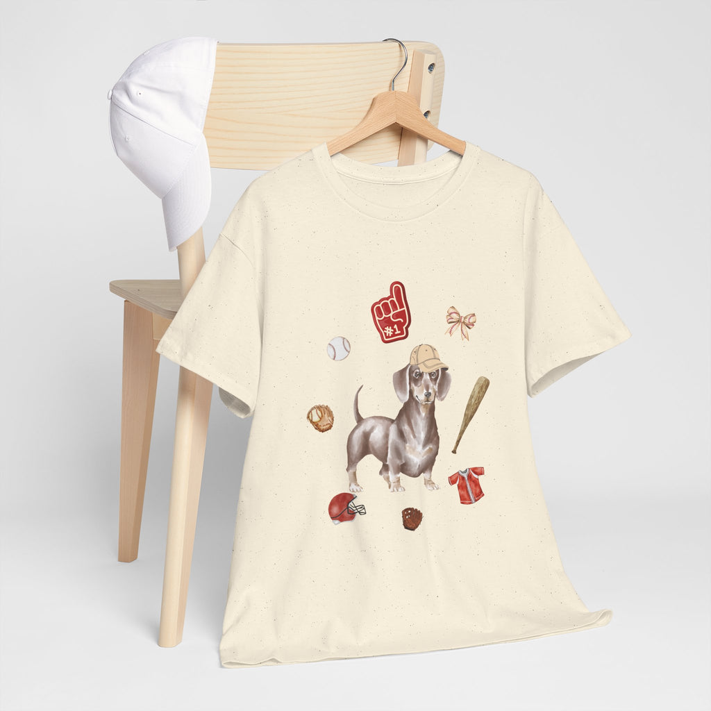 A beige t-shirt hanging on a chair, featuring a graphic design of a dog in a baseball hat surrounded by a circle of balls, bats, and helmets.