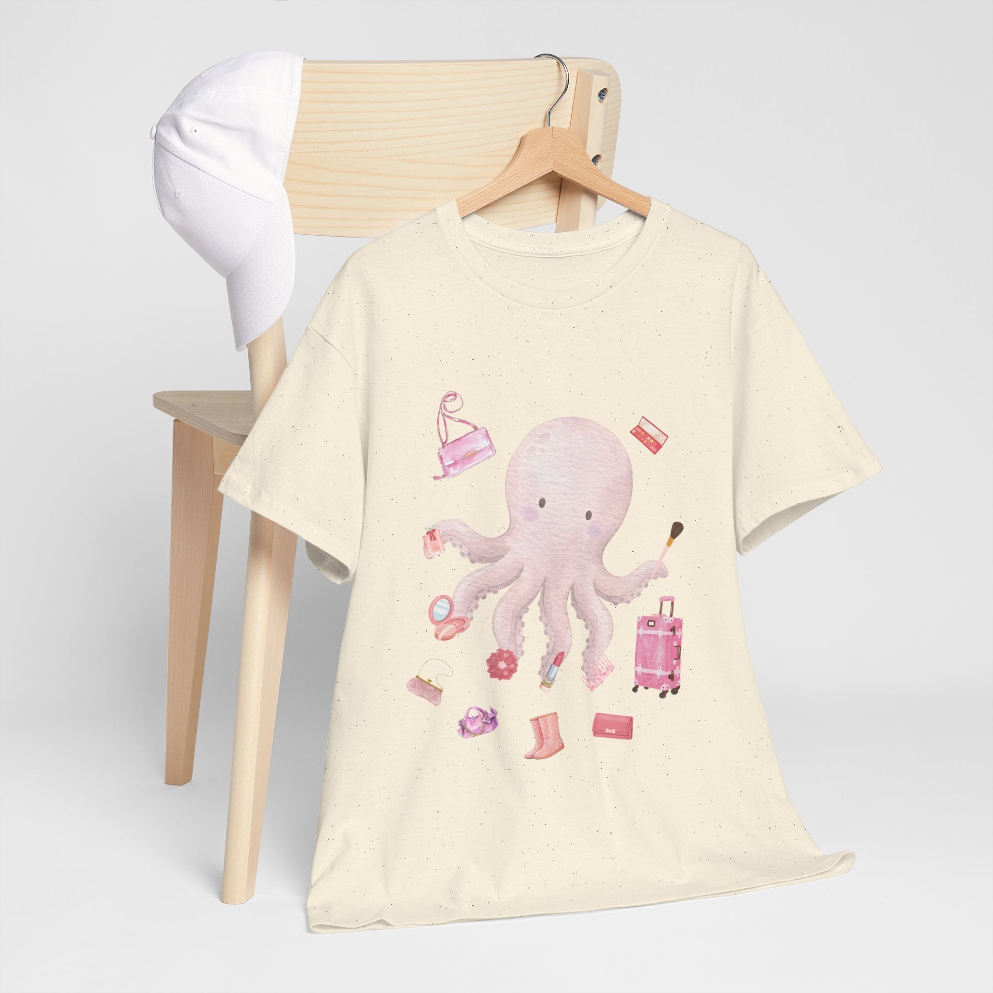 A beige t-shirt hanging on a chair, featuring a graphic design of a pink octopus multitasking its tentacles to apply lipstick, brush on makeup, and juggle beauty accessories.