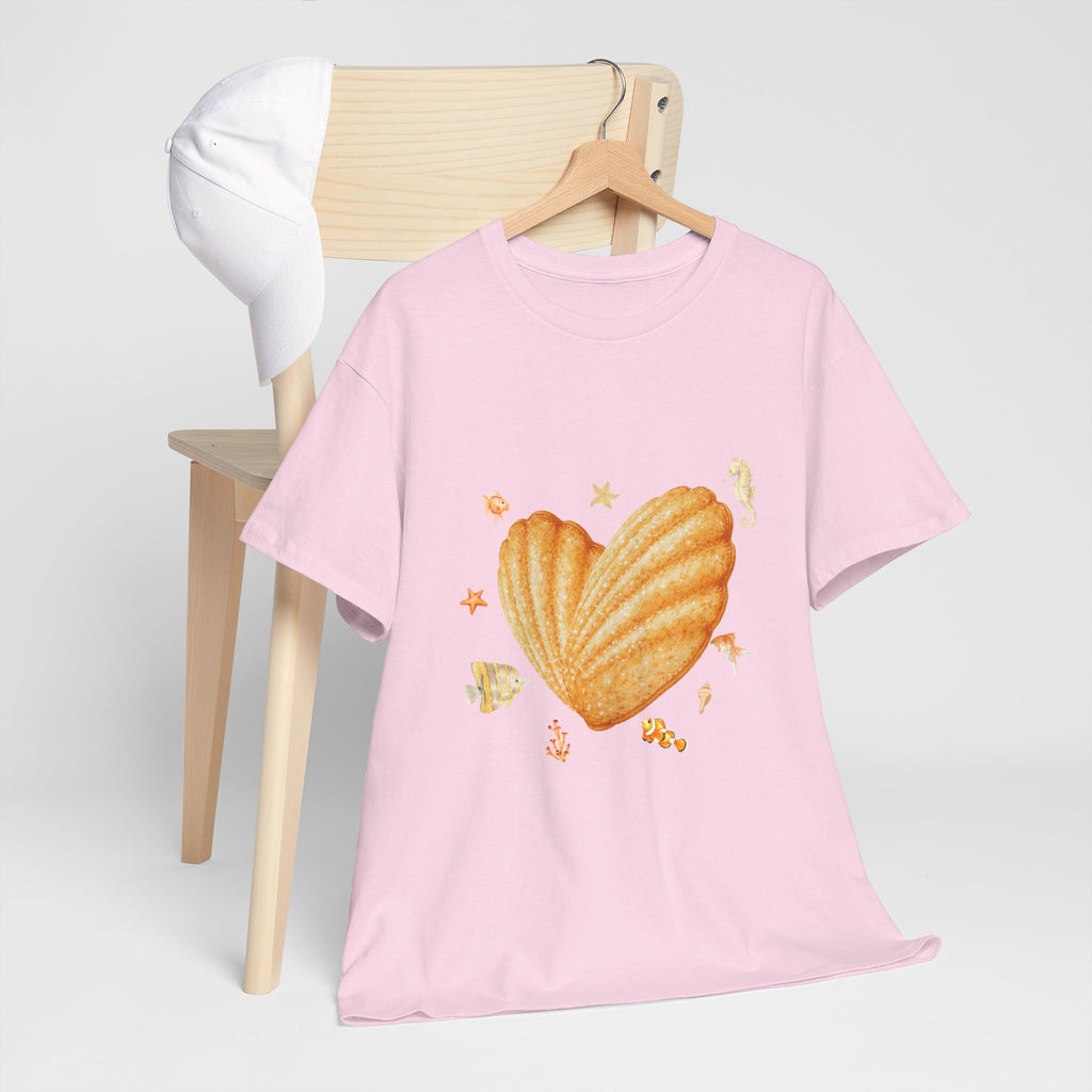 A pink t-shirt hanging on a chair, featuring a graphic design of a heart-shaped madeleine surrounded by yellow tropical fish, starfish, seahorses, and coral.