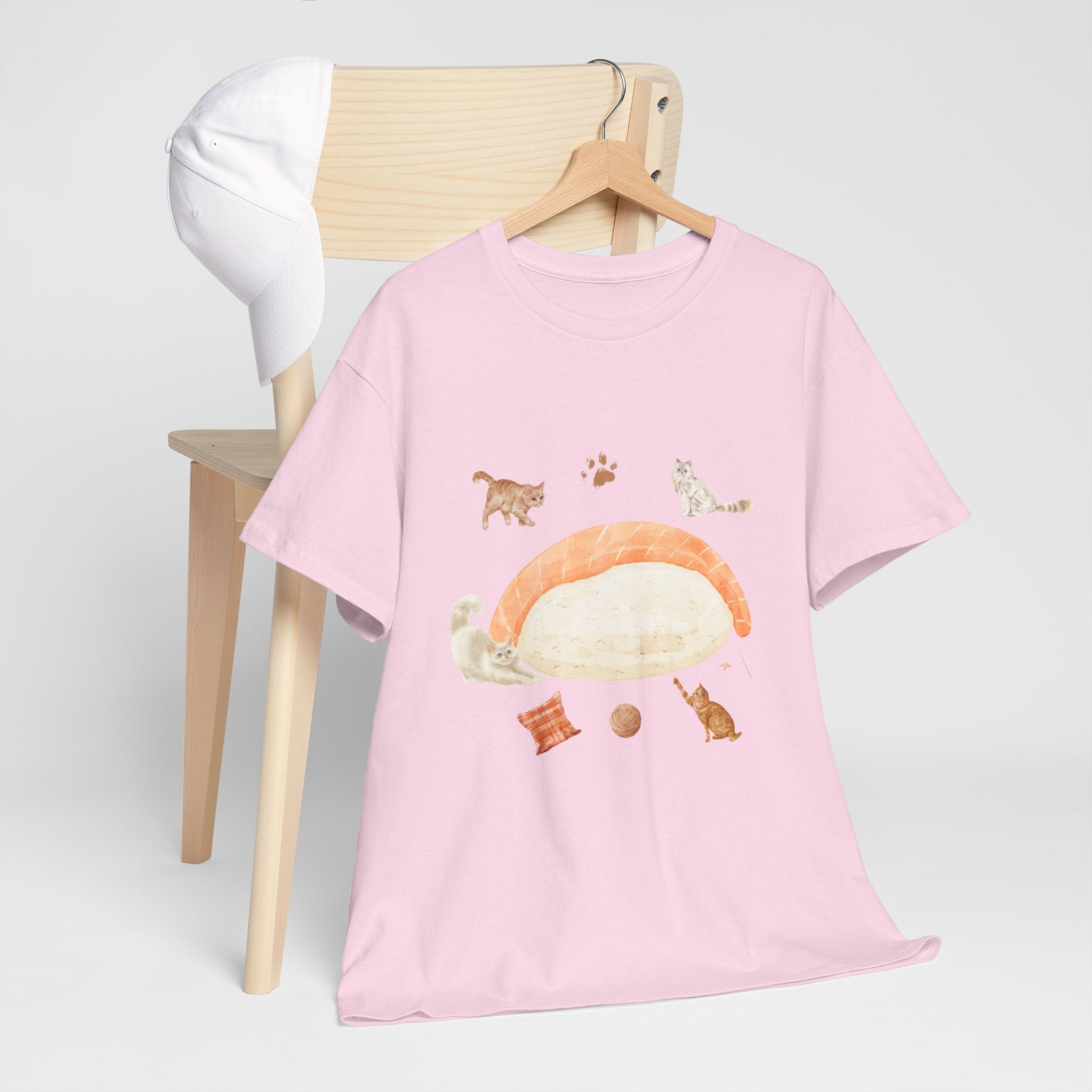 A pink t-shirt hanging on a chair, featuring a graphic design of a piece of nigiri sushi surrounded by a circle of cats, yarn balls, and cushions.