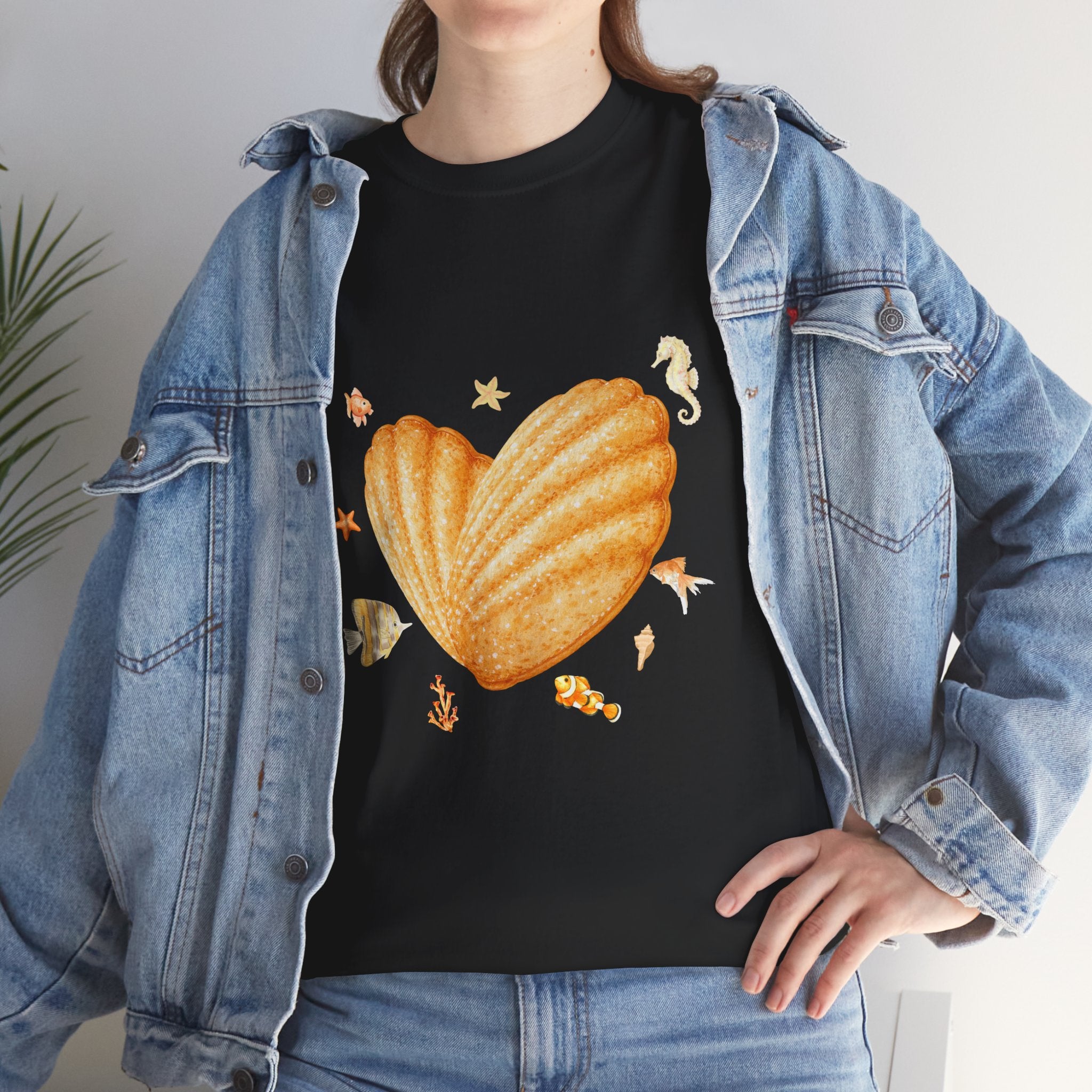 A girl wearing a denim jacket over a black t-shirt featuring a colorful design of a heart-shaped madeleine surrounded by yellow tropical fish, starfish, seahorses, and coral.