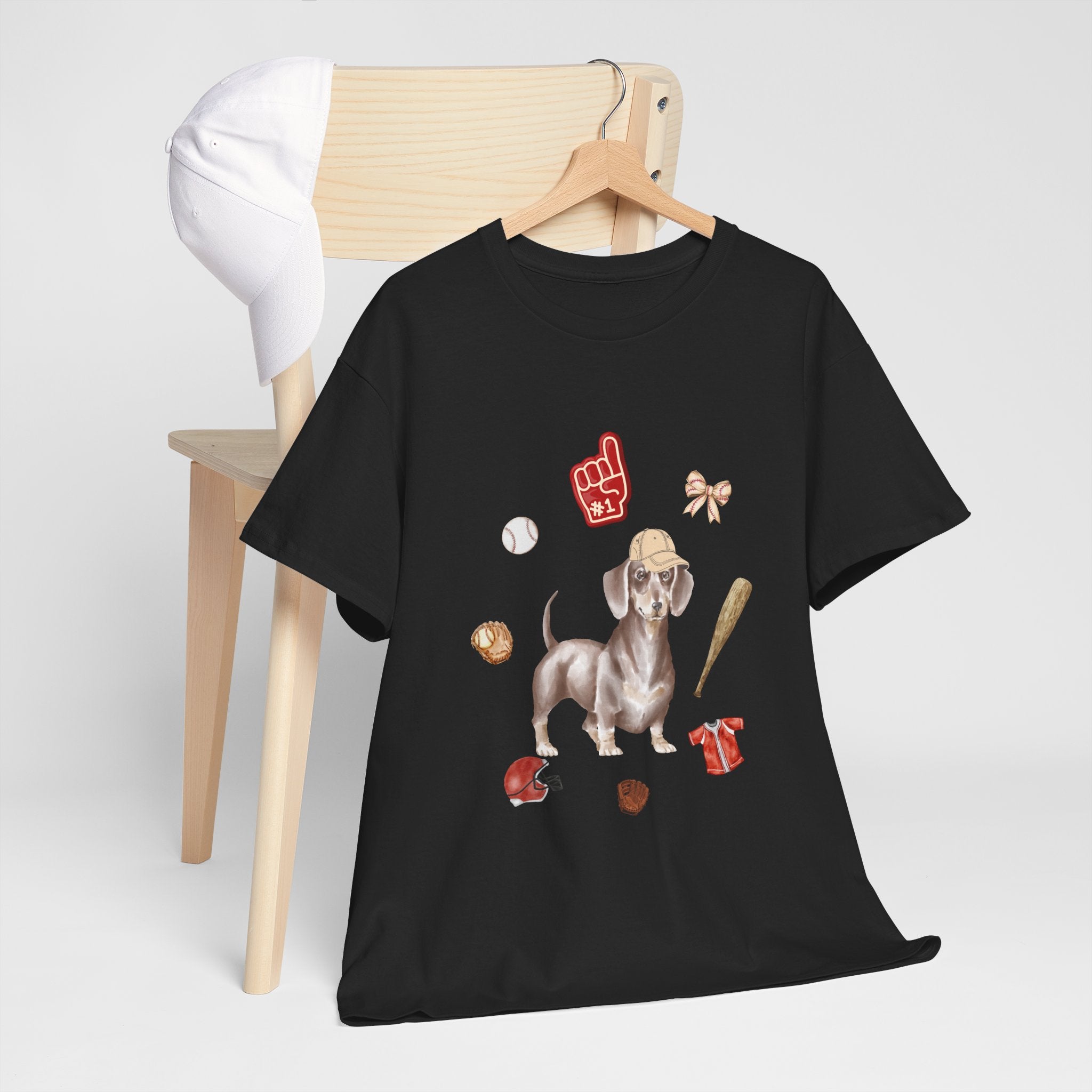 A black t-shirt hanging on a chair, featuring a graphic design of a dog in a baseball hat surrounded by a circle of balls, bats, and helmets.