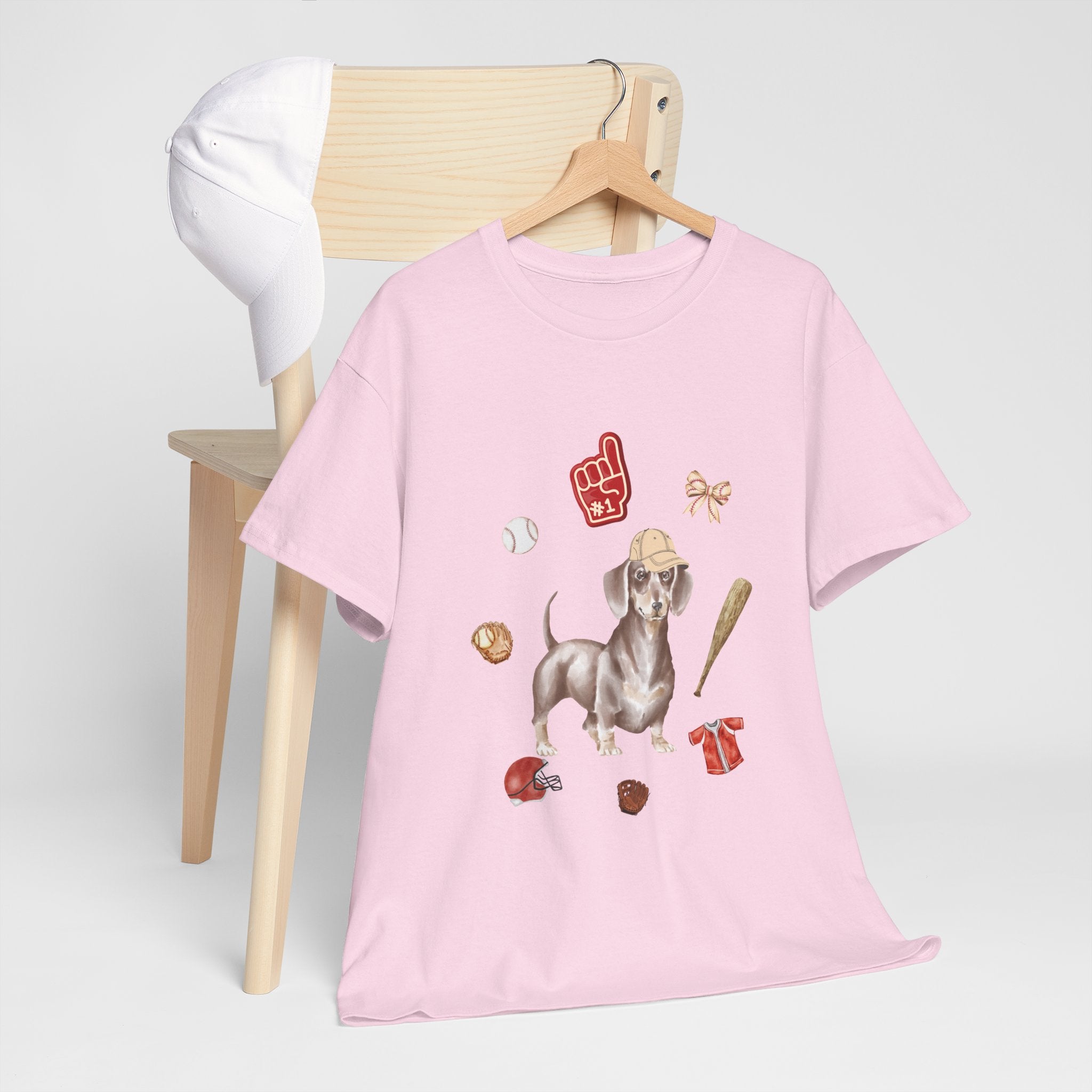 A pink t-shirt hanging on a chair, featuring a graphic design of a dog in a baseball hat surrounded by a circle of balls, bats, and helmets.