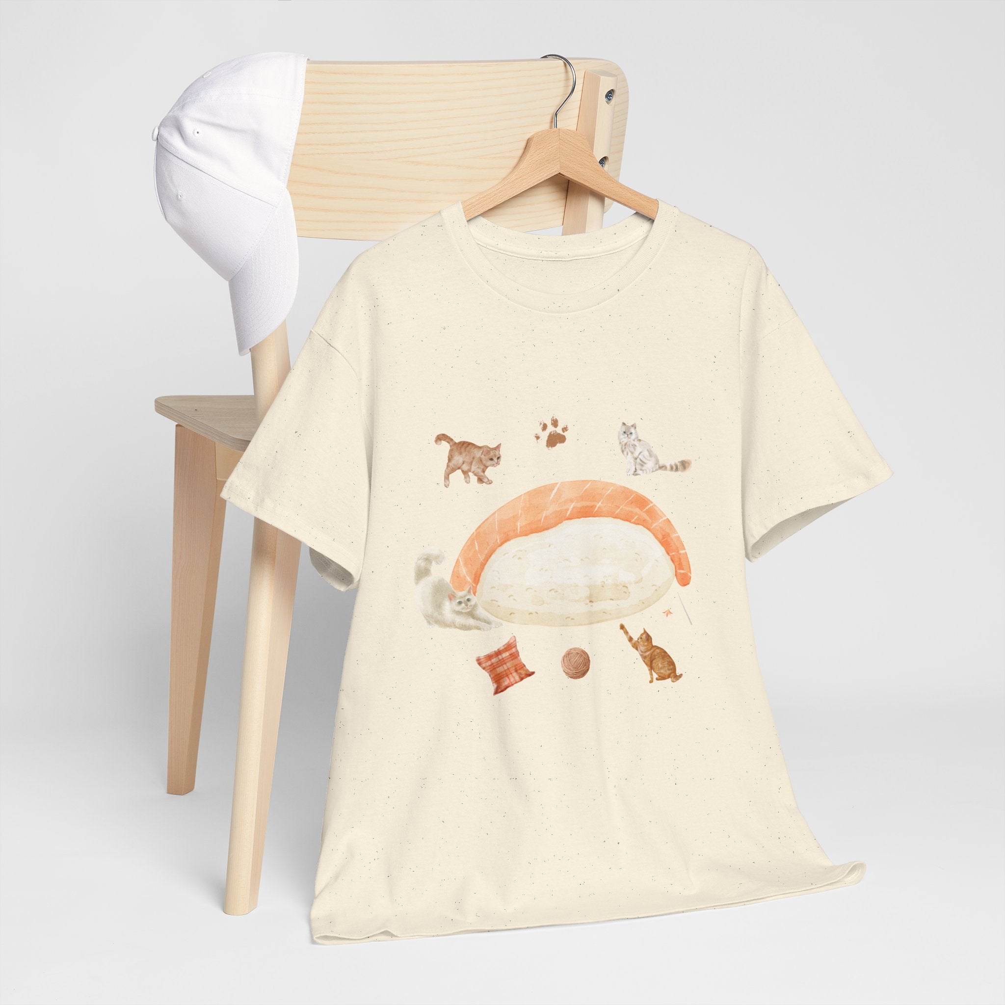 A beige t-shirt hanging on a chair, featuring a graphic design of a piece of nigiri sushi surrounded by a circle of cats, yarn balls, and cushions.