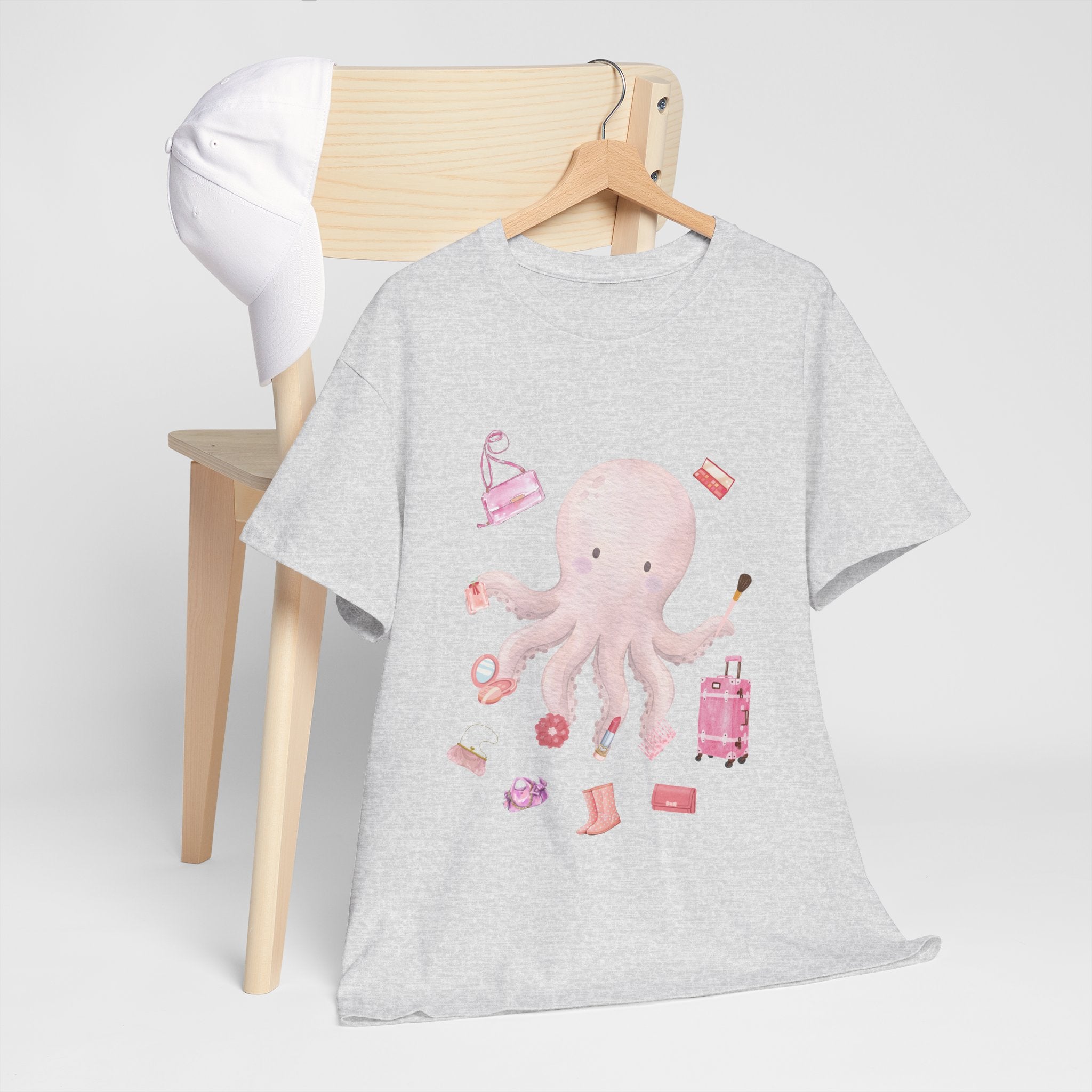 A grey t-shirt hanging on a chair, featuring a graphic design of a pink octopus multitasking its tentacles to apply lipstick, brush on makeup, and juggle beauty accessories.