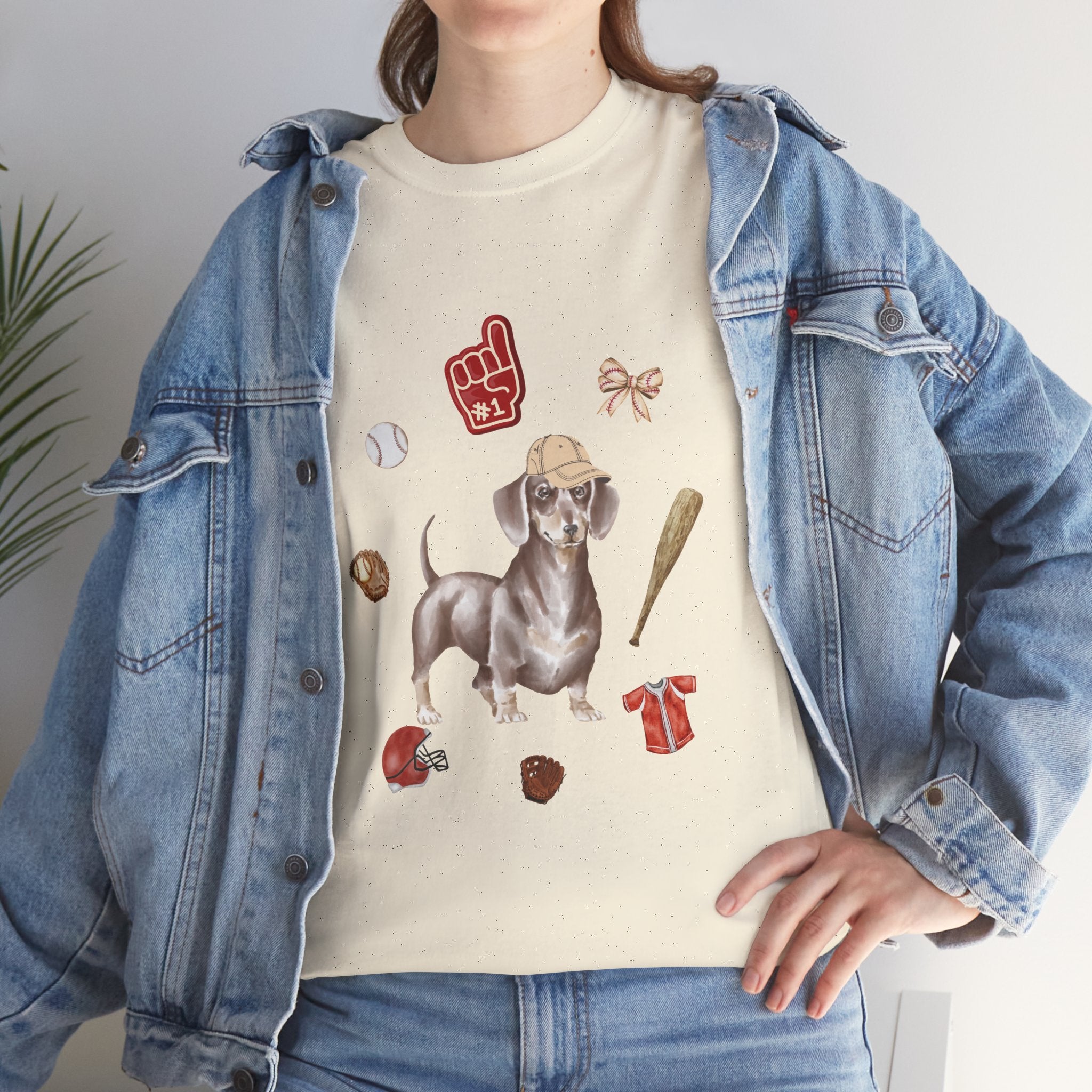 A girl wearing a denim jacket over a beige t-shirt featuring a colorful design of a dog in a baseball hat surrounded by a circle of balls, bats, and helmets.