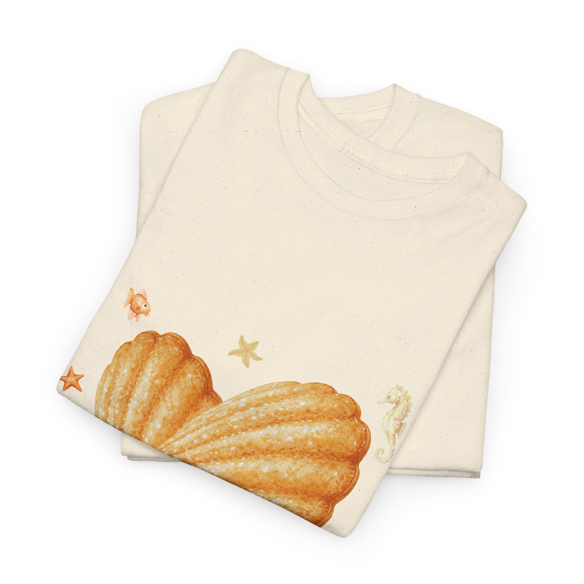 A folded beige t-shirt featuring a graphic design of a heart-shaped madeleine surrounded by yellow tropical fish, starfish, seahorses, and coral.