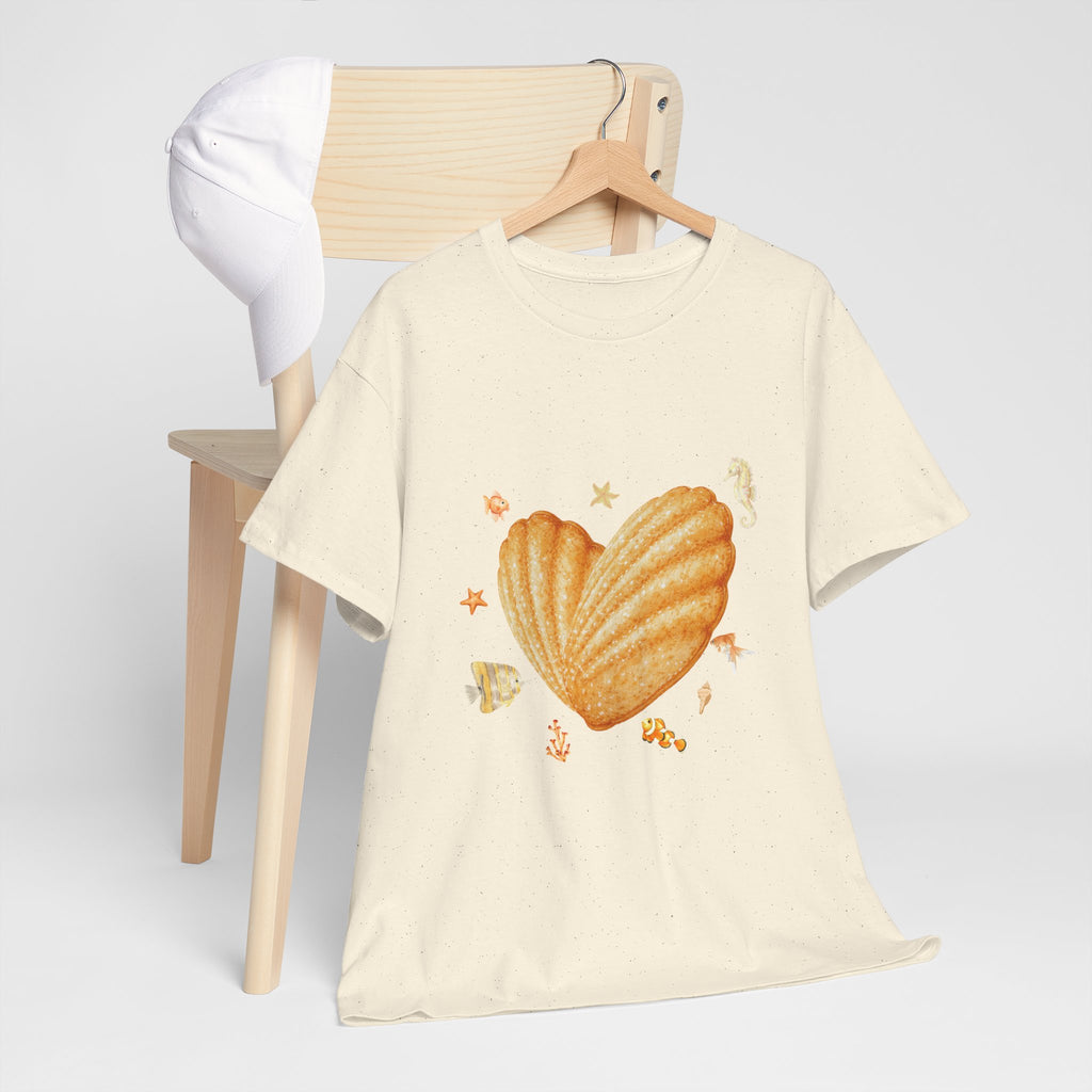 A beige t-shirt hanging on a chair, featuring a graphic design of a heart-shaped madeleine surrounded by yellow tropical fish, starfish, seahorses, and coral.