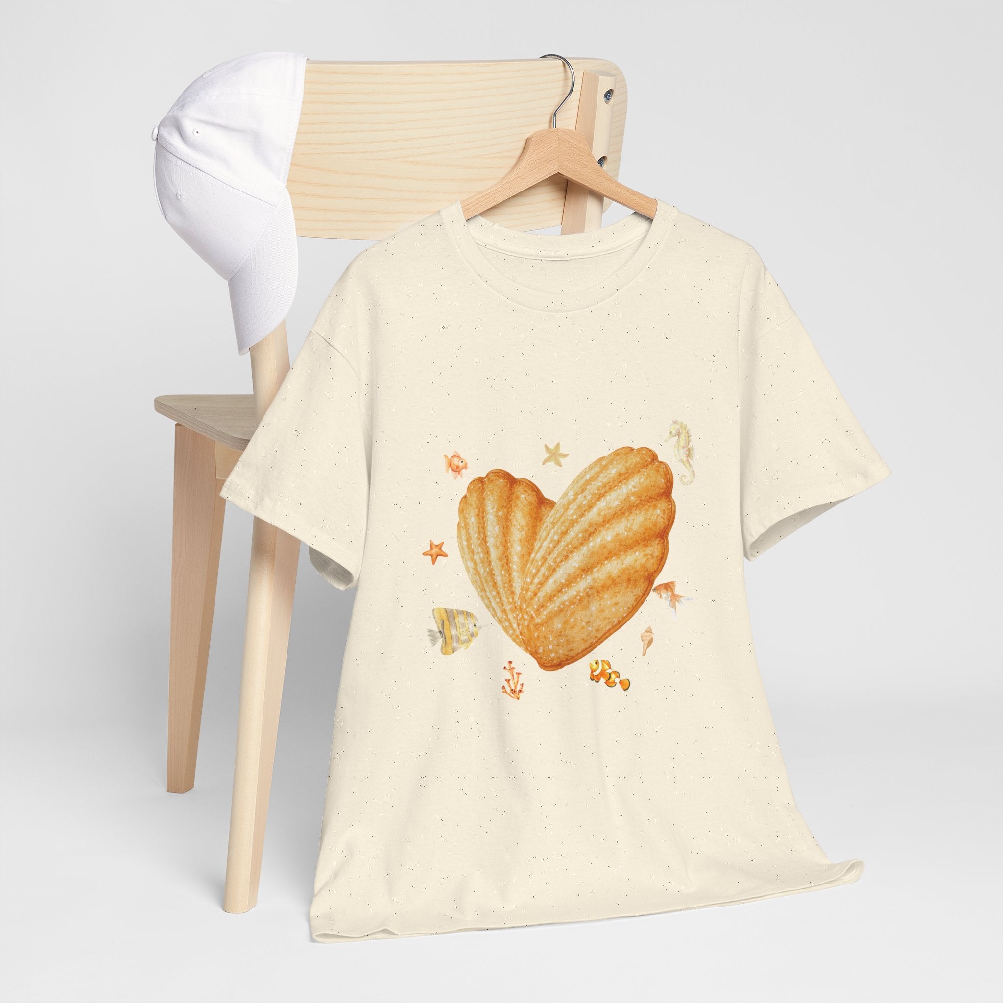 A beige t-shirt hanging on a chair, featuring a graphic design of a heart-shaped madeleine surrounded by yellow tropical fish, starfish, seahorses, and coral.