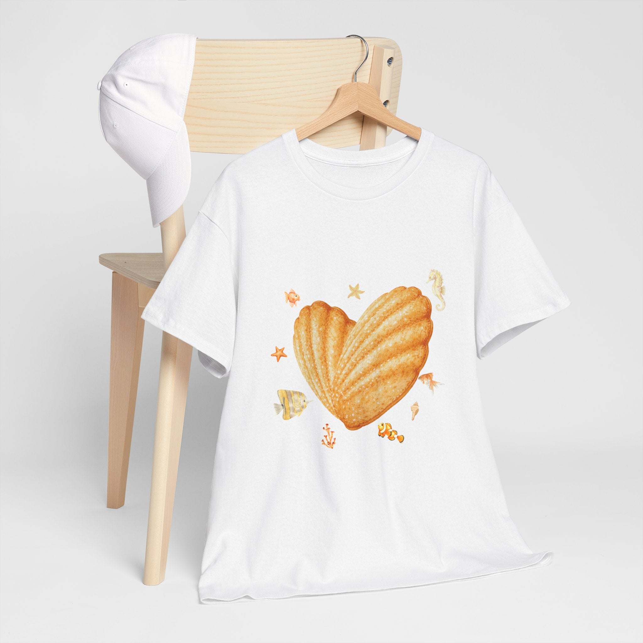 A white t-shirt hanging on a chair, featuring a graphic design of a heart-shaped madeleine surrounded by yellow tropical fish, starfish, seahorses, and coral.