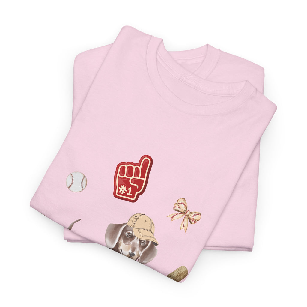 A folded pink t-shirt featuring a graphic design of a dog in a baseball hat surrounded by a circle of balls, bats, and helmets.