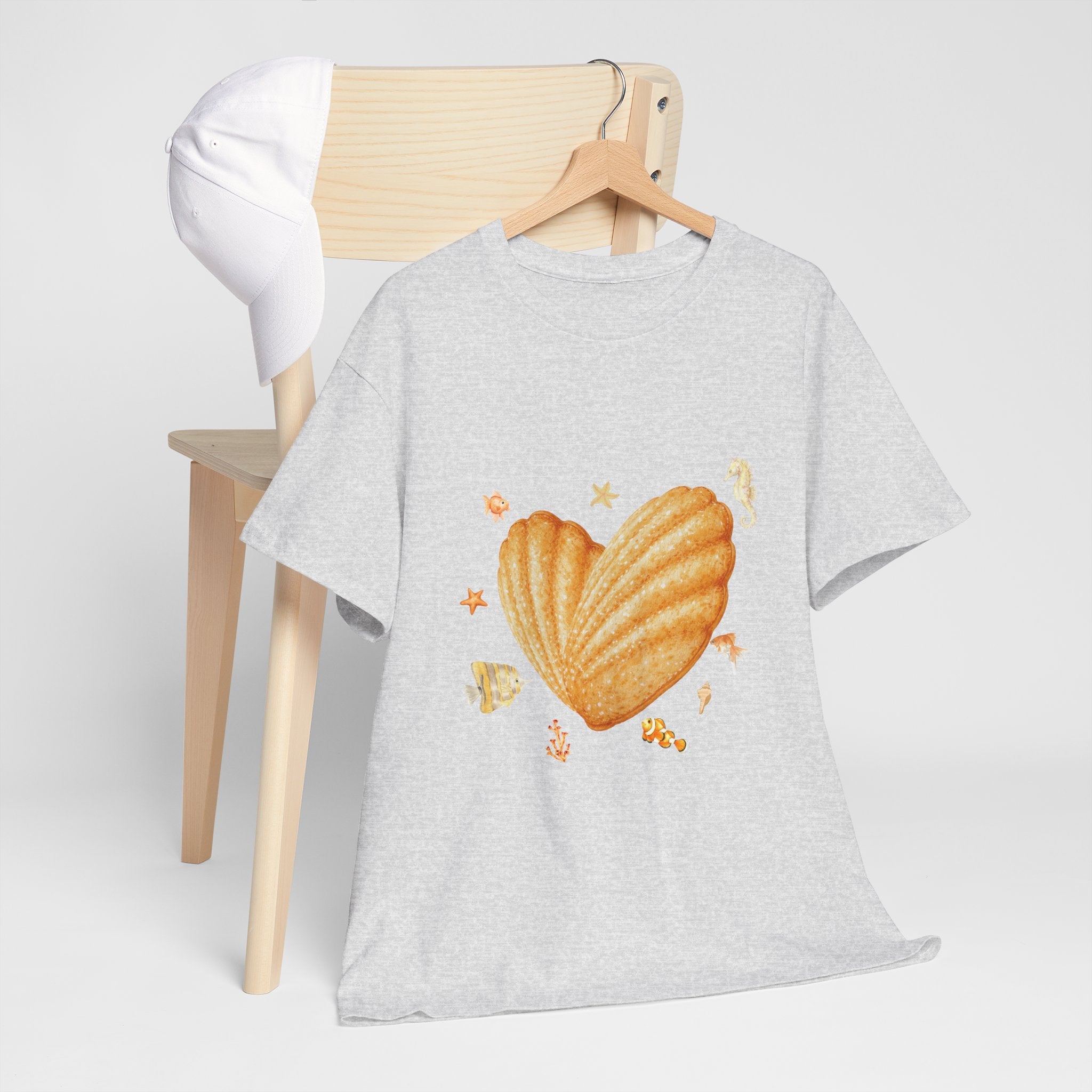 A grey t-shirt hanging on a chair, featuring a graphic design of a heart-shaped madeleine surrounded by yellow tropical fish, starfish, seahorses, and coral.