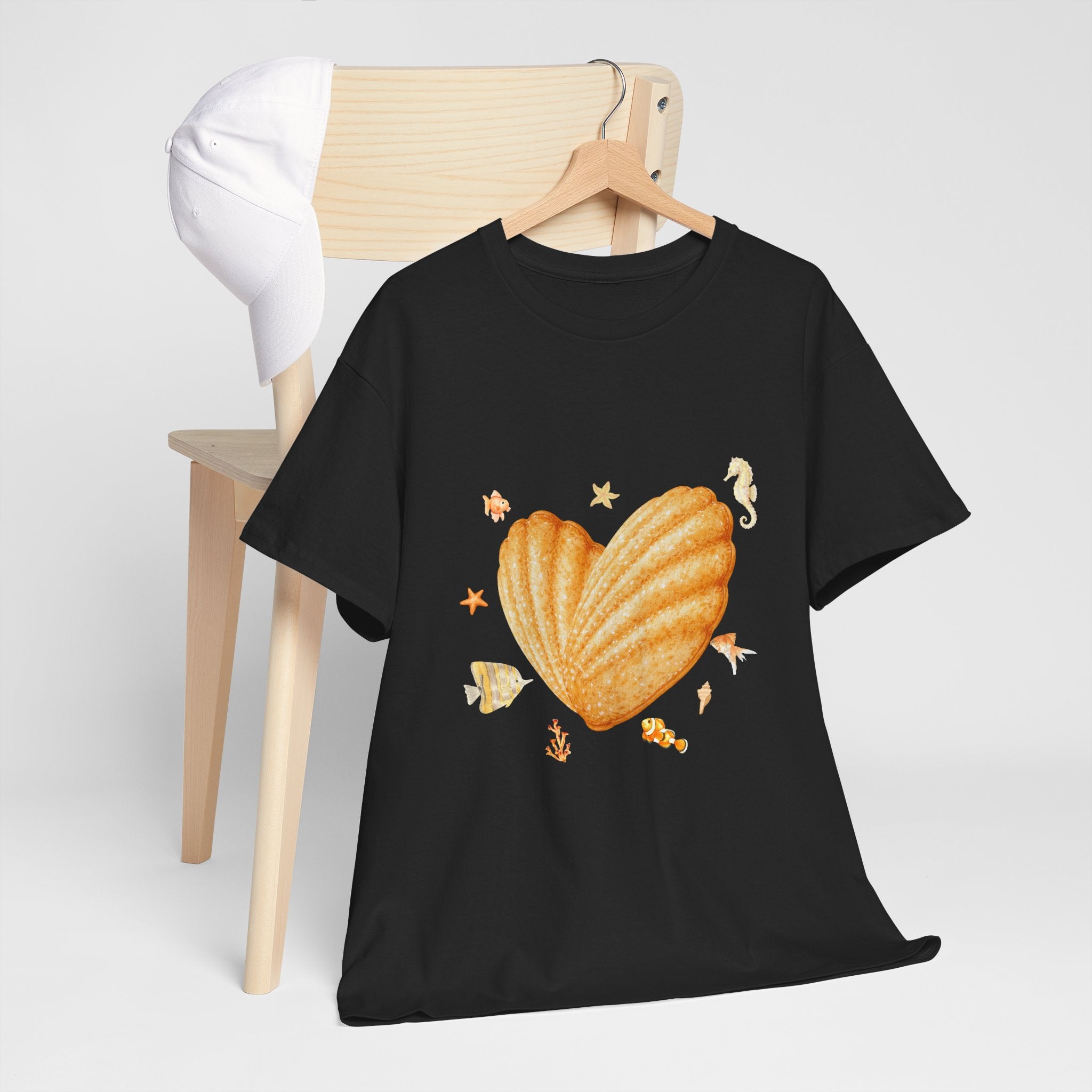 A black t-shirt hanging on a chair, featuring a graphic design of a heart-shaped madeleine surrounded by yellow tropical fish, starfish, seahorses, and coral.