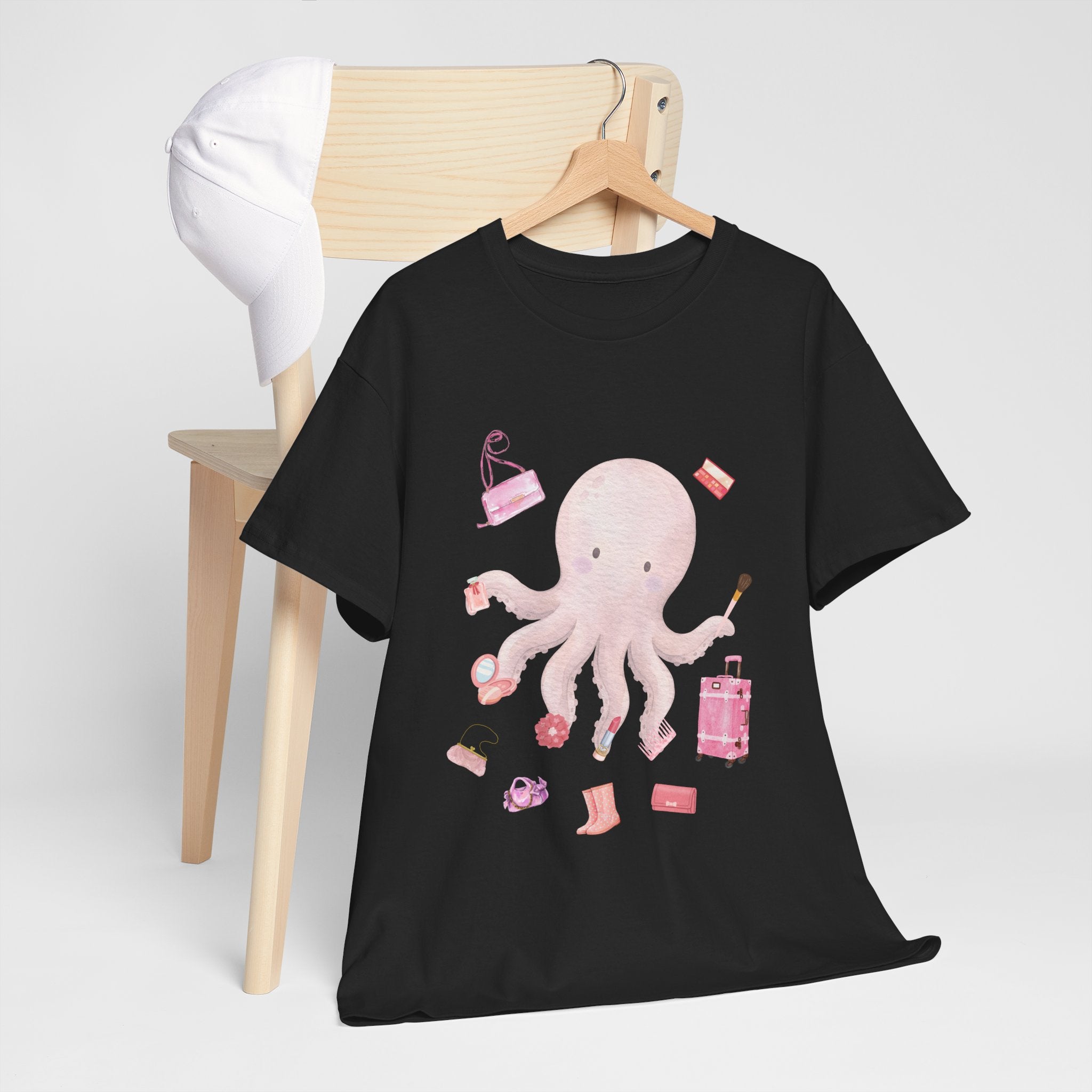 A black t-shirt hanging on a chair, featuring a graphic design of a pink octopus multitasking its tentacles to apply lipstick, brush on makeup, and juggle beauty accessories.